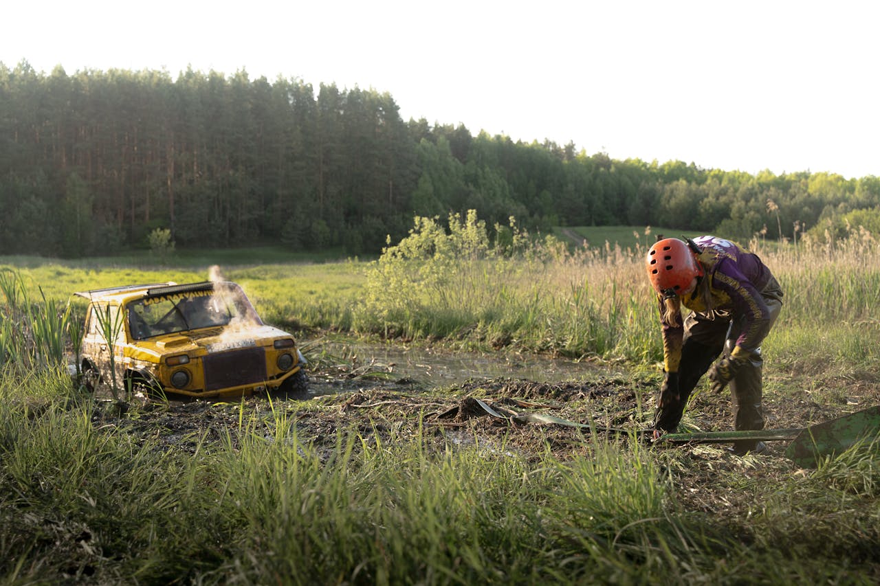 contact-img Person in helmet checks a stuck yellow off-road vehicle in a muddy area.